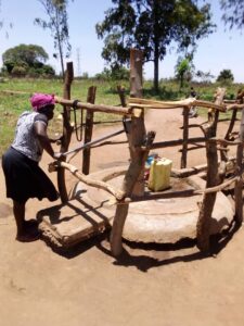 Borehore surrounded by wooden frames, with a woman pumping water into a jerrycan